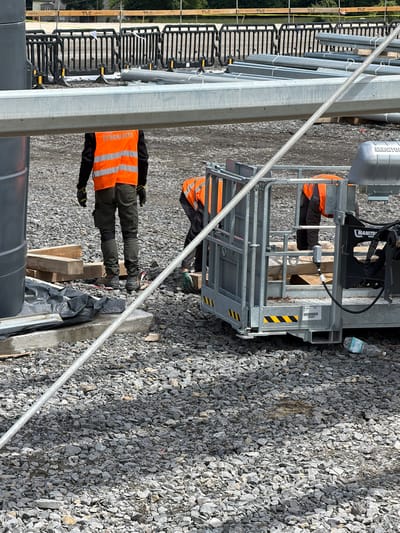 The image depicts a construction site in Jodłownik, Poland, featuring two individuals wearing orange high-visibility vests. One person, facing away from the camera, stands in the foreground wearing olive-green work pants and dark boots. The second individual is partially visible on the right, also in an orange vest, operating a hydraulic lift platform. The platform, branded "MANITOU," has a metal cage and is situated on a gravel surface. Construction materials, including metal beams and pipes, are present in the background, alongside black safety barriers. The environment appears to be outdoors, with a gray, overcast sky suggested by the lighting.