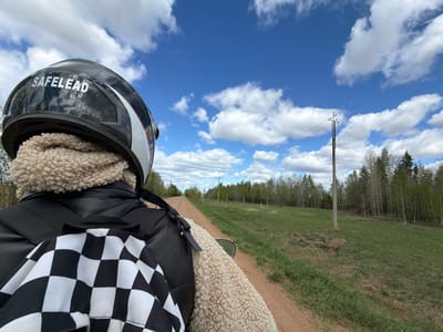 A first-person perspective image shows a person on a motorcycle, facing forward on a dirt road in a rural area. The person is wearing a black "SAFELEAD" brand motorcycle helmet, a beige sherpa-lined jacket, and a black backpack with a black and white checkered pattern hanging from it. The dirt road stretches into the distance, flanked by green grassy fields and rows of young trees. Power lines and poles run alongside the road. The sky above is bright blue with scattered white clouds. Based on the presence of power lines along a rural road and the specific type of vegetation, this image is likely taken in or near the city of Можга, Russia.