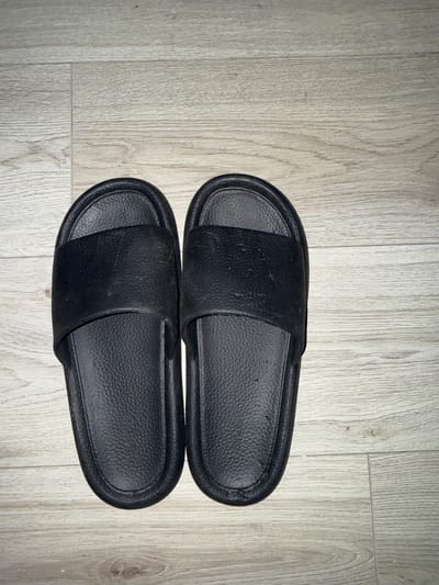 The image shows a pair of black slide sandals resting on a light-colored wooden floor. The sandals are oriented horizontally, with the left sandal slightly above and to the left of the right sandal. Both sandals appear to be made of a textured material. The wooden floor has a visible grain pattern. There are no people or other objects in the image. The scene is indoors, on a floor. The location is Hainstadt, Germany.