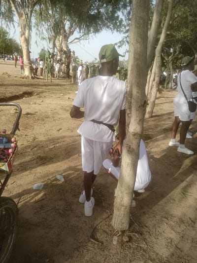 The image captures a scene in the city of Majemu, Nigeria, featuring a group of young people, likely participants in a youth service program given their matching white uniforms and green caps. In the foreground, a young man in a white t-shirt, white shorts, and white sneakers stands with his back to the camera, leaning against a tree trunk. He is wearing a green cap. To his right, another young man, also in white attire and a green cap, is visible from the waist down, wearing a black crossbody bag. Further back, a person in white is crouched near the base of a tree. The environment is dusty with scattered debris and patches of dry vegetation, typical of a semi-arid or rural area. Several trees with large trunks and sparse foliage dominate the background, with more people in similar uniforms milling around under their shade. The overall atmosphere suggests an outdoor gathering or event taking place during the daytime.