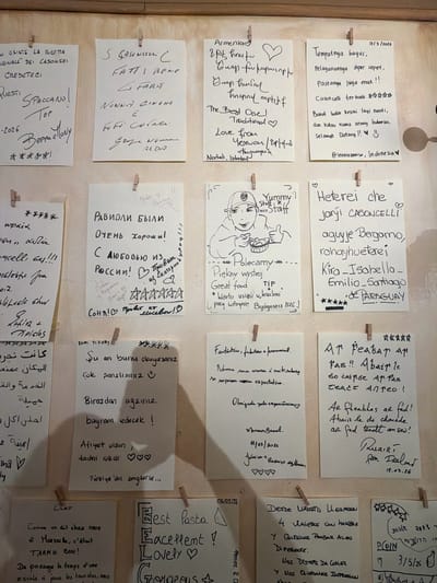 The image shows a close-up, eye-level shot of a wall adorned with numerous handwritten notes, possibly in a restaurant or public space in Mozzo, Italy. The notes are of various sizes, shapes, and colors, predominantly white paper, and are attached to the wall with small wooden clothespins. The handwriting styles are diverse, ranging from neat and cursive to more casual scribbles. Some notes contain drawings, such as a sketch of a person eating. The notes appear to be reviews or messages from patrons, written in multiple languages including Italian, Armenian, Russian, Turkish, Portuguese, French, and English. Some notes include dates and star ratings. The overall impression is a collection of personal testimonials and expressions of appreciation. The lighting appears to be ambient, and the background is a neutral, textured wall.