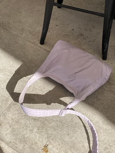 A light purple fabric bag is lying on a light grey tiled ground in Megalochori, Greece. The bag, made of a soft, slightly wrinkled material, has an extended strap across the tiles. To the upper right, two legs of a black metal chair or stool are visible. A small, dried brown leaf rests on the ground in the lower left. Strong, direct sunlight illuminates the scene, casting sharp shadows, including a large, irregular shadow on the left and shadows from the bag and chair legs. No individuals are present or performing any actions.