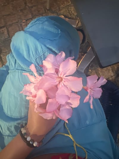 A person is holding a bouquet of pink flowers against a backdrop of a blue garment, likely a dress or skirt. The person's arm is visible, adorned with a bracelet composed of various beads. The background consists of a textured surface, resembling cobblestones or brickwork, suggesting an outdoor setting. A dark, flat surface, possibly a table, is partially visible to the right, along with a dark woven texture that could be a chair. The lighting appears dim, casting shadows and contributing to a slightly muted color palette. The image is a close-up, focusing on the flowers and the person's arm and clothing. The exact location within Noyon, France, cannot be determined from the visual information alone.