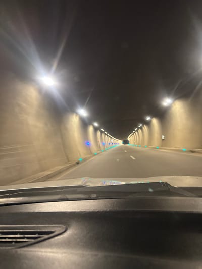 A long, brightly lit tunnel recedes into the distance, vanishing into a dark point. Lights are arranged on either side of the tunnel, with both bright white and blue lights visible. The image is taken from inside a vehicle, with a portion of the interior visible on the right side. The vehicle's interior is dark gray and features vents. The tunnel is concrete, and the lighting creates a sense of depth and perspective.