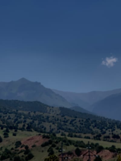 The image depicts a panoramic view of rolling hills and mountains under a clear blue sky. In the foreground, there are patches of green vegetation and some exposed reddish-brown earth, possibly indicating tilled land or a landslide. A few scattered trees dot the landscape. In the mid-ground, a densely wooded hillside rises, with varied shades of green suggesting different types of foliage. In the background, layers of hazy, blue-hued mountains recede into the distance, their peaks softened by atmospheric perspective. A small, wispy white cloud is visible in the upper right portion of the sky. The overall scene conveys a sense of natural, mountainous terrain with signs of human presence in the form of cultivated or disturbed land. The location is specified as Dardar, Uzbekistan.