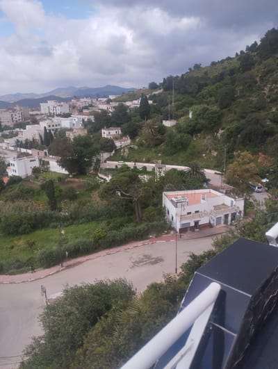The image shows a view overlooking a neighborhood in Tétouan, Morocco, characterized by white buildings with reddish roofs and walls, scattered across a hilly landscape. The foreground features a road with sidewalks, streetlights, and green vegetation. A structure resembling a balcony with a black and white railing is visible in the lower right corner. In the midground, there are white buildings along the road, surrounded by trees and green spaces. In the background, a range of mountains are visible under a cloudy sky. A person is walking along the sidewalk in the lower left area.