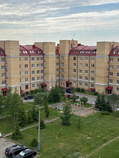 An aerial view captures a modern residential apartment complex in the city of Kirishi, Russia. The building, constructed with light yellow bricks and featuring distinctive red, mansard-style roofs with dormer windows, forms a U-shape around a central courtyard. The courtyard is a vibrant green space with mature trees and a children's playground equipped with swings and climbing structures. Several vehicles are parked on asphalt surfaces surrounding the complex and within the courtyard. The sky above is filled with a dramatic pattern of grey and white clouds, suggesting a cloudy or overcast day. A single person is visible walking in the courtyard.