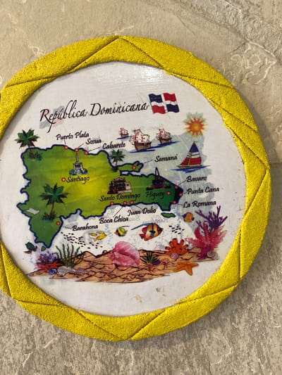 The image is a circular decorative piece, likely a coaster or a small plaque, featuring a map of the Dominican Republic. The map is colorful and cartoonish, with illustrations of palm trees, beaches, sailboats, historical ships, and underwater life like fish and coral. Several cities on the island are marked with red dots and labeled, including Puerto Plata, Sosua, Cabarete, Santiago, Samana, Santo Domingo, Higüey, Juan Dolio, Boca Chica, Barahona, Bavaro, Punta Cana, and La Romana. The Dominican flag is also depicted. The circular piece has a textured yellow border. The background appears to be a textured, light-colored surface, possibly a wall or table. There is no indication of Yokohama, Japan, within the image.