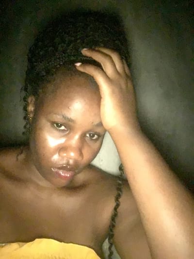 A woman with dark skin and dark, curly hair lies on her side, looking up at the camera. Her right hand rests on her head. Her skin has a glossy appearance. She has a small earring in her left ear. A yellow cloth or blanket is visible on her right side. The background appears dark, possibly a wall or floor. The image is taken from a low angle.