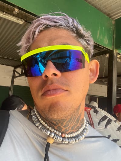 A person with light purple hair and a neck tattoo wears bright yellow-rimmed blue reflective sunglasses. They are wearing a gray shirt and multiple strands of beaded necklaces, including pearl and silver-colored beads. The background includes green paneling and glimpses of a patterned shirt and a red cap. The reflection in the sunglasses appears to show a bus or van.