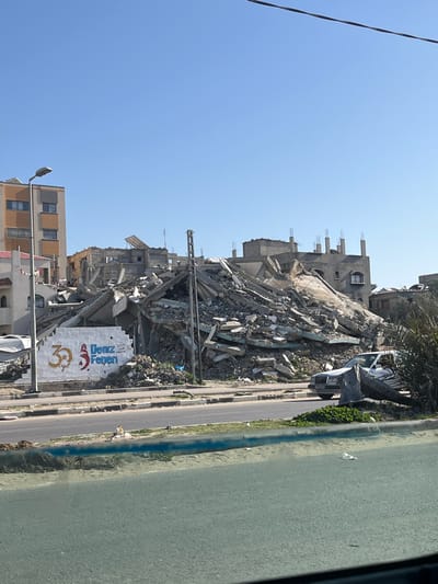 The image presents a somber scene of widespread destruction, likely the aftermath of a major disaster such as an earthquake. Under a bright and clear blue sky, massive piles of concrete, rebar, and other building debris dominate the central and right portions of the frame, indicating the complete collapse of multiple multi-story buildings. Some partially standing structures are visible in the background, exhibiting severe damage with exposed frameworks and broken walls.

In the foreground, a paved road or street runs along the bottom of the image, flanked by remnants of destruction. A dark-colored car, possibly a sedan, is visible on the roadside, partly obscured by a small palm tree and debris. Further down the road, another vehicle is partially visible.

A prominent feature in the lower right foreground is a large white billboard or sign. The text "Deniz Feneri" is clearly visible in blue lettering, accompanied by a logo that includes a red crescent moon and star, and the number "30". "Deniz Feneri" is a well-known Turkish humanitarian aid organization, suggesting the scene is located in Turkey or a Turkish-speaking area.

On the far left, a multi-story building appears relatively more intact compared to the surrounding devastation. A streetlight on a pole stands next to this building. A dark line, possibly a power line or utility cable, stretches across the upper left corner of the clear blue sky.

The bright daylight and clear sky offer a stark contrast to the grim reality of the collapsed buildings and rubble below, conveying a powerful sense of loss and devastation. The presence of the aid organization's sign indicates the aftermath is being addressed by humanitarian efforts.