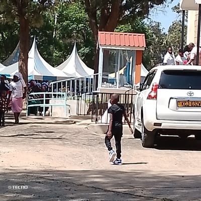 The scene depicts an outdoor gathering in Machakos Central ward, Kenya, under a clear sky. In the foreground, a young boy, dressed in a black t-shirt, black pants, and blue and white sneakers, walks from left to right, with his back partially visible. To the left, a woman in a white top, pink skirt, and white head covering walks towards the left, accompanied by a child in a patterned shirt. In the midground, a white Toyota Land Cruiser Prado, bearing license plate KCJ 032W, is parked on the right. Adjacent to the vehicle is a small shrine-like structure featuring a red tiled roof and a glass enclosure containing a statue. Behind a light blue metal railing, several white peaked tents are erected, beneath and around which numerous individuals are gathered, some seated. A light-colored building is partially visible on the far right, with additional people standing near its entrance. The environment is paved with areas of dirt and features mature trees in the background.