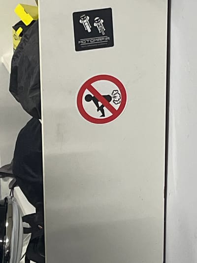 The image shows a portion of a white surface, likely a cabinet or wall, bearing two stickers. One sticker is rectangular and black, featuring two stylized images of motorcycles and the word "MOTOMAFIJA". The other sticker is round, with a red circle and a diagonal line through it, depicting a stick figure riding a scooter with visible exhaust, indicating a prohibition of scooters emitting exhaust. Below the white surface, there are dark-colored soft materials, possibly bags or clothes, and a portion of a chrome object. The setting appears to be indoors. There is no evidence to suggest the location of this image is in Grīnvalti, Latvia.