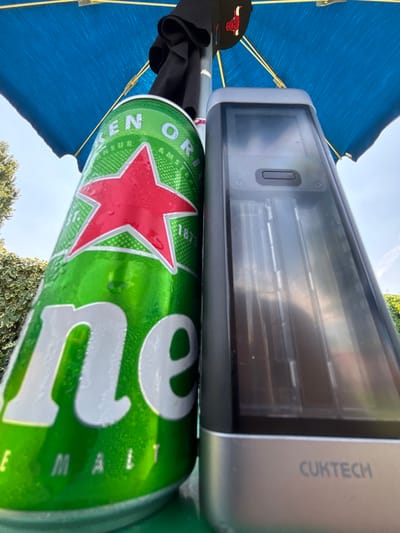 The media file shows an outdoor scene, possibly a backyard or patio, as suggested by the foliage in the background and the bright, clear sky indicating daytime. The primary objects in focus are a can of Heineken beer and a CUKTECH electronic device, likely a portable charger or battery pack. The Heineken can is green with the iconic red star and white lettering, and it appears to be cold and slightly sweating. The CUKTECH device is silver and rectangular with a screen and a button visible. It is positioned above the beer can.
The scene is framed by a blue umbrella, providing shade. Partially visible beneath the umbrella and next to the beer can is a black baseball cap with a red bull logo. The lighting suggests it is a sunny day, likely midday. The overall impression is one of relaxation and leisure, perhaps someone enjoying a drink and their device outdoors. There is no clear indication of people, but the items suggest a single person's presence. The text visible includes "HEINEKEN" and "CUKTECH." The location context of Vicenza, Italy, doesn't add specific visual details to this particular frame but suggests an Italian setting.