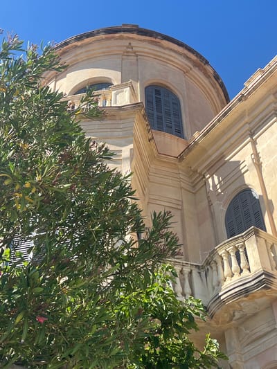 The image shows a building facade with baroque architectural elements, including arched windows with closed shutters, decorative moldings, and a balustrade. The building is light-colored, possibly sandstone or a similar material. A dense green shrub with long, narrow leaves is in the foreground, partially obscuring the view of the building. The sky is clear and blue in the background. The location is indicated as Sant Pol De Mar, Spain, but this cannot be confirmed based solely on the visual information.