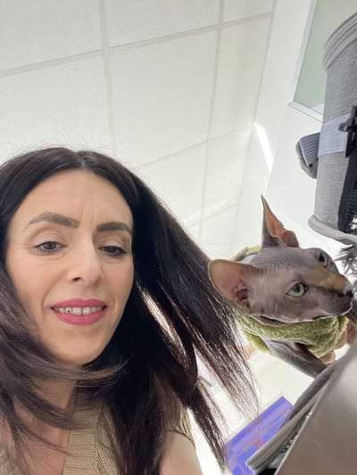 This image is a low-angle selfie featuring a smiling woman and a Sphynx cat in an indoor setting, located in Wasserliesch, Germany. The woman, with long dark hair and red lipstick, is positioned in the bottom right, smiling brightly towards the camera. Her face is angled upwards, and some wrinkles are visible on her forehead. The Sphynx cat, distinctive for its hairless appearance, large ears, and green eyes, is prominently featured in the upper middle section, looking down towards the camera. It is wearing a light green knitted sweater or scarf.

In the background, a white drop ceiling with square grid panels is visible, suggesting an office, clinic, or similar commercial indoor environment. A gray pet carrier or bag is partially visible in the upper left corner of the frame, and another dark object is seen in the upper right. The scene conveys a casual moment, possibly an owner interacting with their pet. No specific time of day is discernible, and no text is visible within the image. The woman appears happy and relaxed, while the cat has a curious or neutral expression.