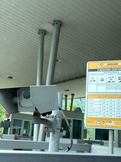 The image shows a close-up of a toll booth camera and a price list sign at a toll plaza. The camera is a grey box with a blue lens and a bright LED light array. It is mounted on a metal pole. The price list sign is yellow and white and lists toll prices for different vehicle classes and locations. The background shows the roof structure of the toll plaza, supported by grey pillars. In the distance, a toll booth building with glass windows is visible, along with some greenery. The sign displays "AC JP Autoceste FB" which refers to the company "Autoceste Federacije Bosne i Hercegovine" (Motorways of the Federation of Bosnia and Herzegovina). The location "Jehovina" is indicated on the sign with a specific toll route. The overall scene depicts a functional toll collection point on a road.