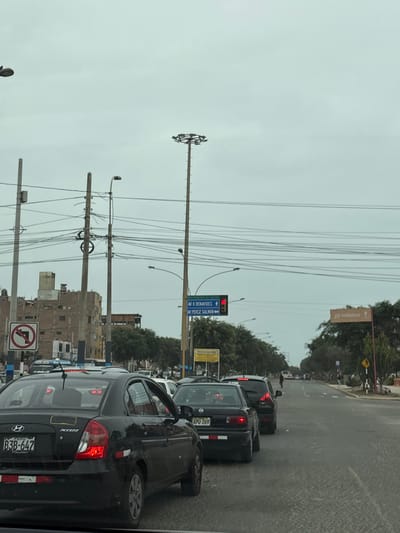 The image depicts a traffic scene in Callao, Peru, on a cloudy day. Several cars are stopped in traffic on a paved road. In the foreground, a black Hyundai Accent is visible, with its license plate reading "PERU B3B-647". Behind it, other cars are also waiting. Overhead power lines crisscross the sky, supported by tall utility poles. Road signs are present, including a blue directional sign indicating "AV 6 BENAVIDES" and "AV PEREZ SALMON", and a red circular sign with a diagonal line indicating "no left turn". In the background, buildings and trees line the side of the road. The overall impression is one of urban congestion under an overcast sky.