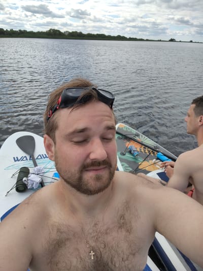 Two shirtless men are on paddleboards on a body of water. The man in the foreground is facing the camera with his eyes closed and has a beard and goggles on his forehead. He is wearing a gold necklace with a cross pendant. In the background, the second man is facing away from the camera. Both men are on white paddleboards, and the one in the foreground has a paddle and a green waterproof speaker. The water is calm, and there is a shoreline with trees in the distance. The sky is overcast with scattered clouds. The image was taken in Shchiglytsy, Russia.