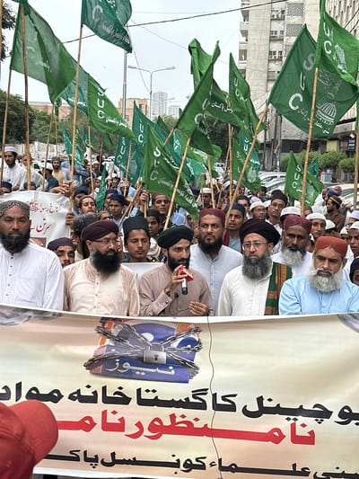 This media file captures a protest or demonstration in Karachi, Pakistan. A large group of men, many wearing traditional Islamic attire and headwear, are visible, holding numerous green flags on bamboo poles. The flags bear the symbol "SUC" and Islamic imagery, along with text in Urdu. In the background, a tall banner with Urdu text and a graphic depicting a locked chain is prominently displayed. The men appear to be engaged in a vocal gathering, with some holding microphones, suggesting a public address or rally. The setting appears to be an urban street, with buildings and power lines visible in the distance, indicating a daytime event under cloudy or overcast conditions. The overall mood seems to be one of solidarity and possibly protest, given the context of the flags and banner. The visible text on the flags includes "کونسل پاکستان" (Council Pakistan) and "گستاخ رسول" (Insolent Prophet). The banner contains phrases like "سُنی علماء کا پاکستان" (Sunni Scholars' Pakistan) and "بوچینل کا گستاخانه مواد نا منظور" (Bochannel's insolent material unacceptable).