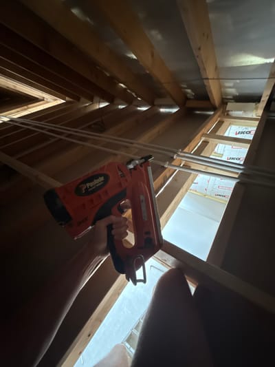 This image depicts an unfinished interior space, likely an attic or upper floor under construction, located in Skipsea, United Kingdom. The precise address or specific type of building within Skipsea is not discernible.
The central subject is a human right hand, with fair skin, grasping an orange and black Paslode IM350+ cordless gas framing nailer. The nail gun is held with the barrel pointed upwards and to the left, and the trigger mechanism is visible. The hand appears to be positioned to operate the tool.
The environment consists of exposed wooden structural elements, including rafters and ceiling joists, indicating ongoing construction or renovation. Between some of the wooden beams, a silver, reflective foil material is visible, consistent with insulation or a vapor barrier. Below this, towards the right side of the frame, multiple sections of white rigid insulation board with red lettering, including "JKO THERM," are visible. Several white plastic pipes, likely for plumbing or heating, run horizontally across the middle ground, secured to the wooden framework. Natural light illuminates the lower section of the frame through an opening in the structure, revealing a brightly lit area below, possibly an adjacent room or an exterior opening. The overall lighting suggests an indoor setting with some external light sources.