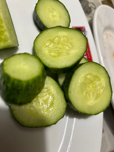 The image shows a close-up view of sliced cucumbers on a white plate. The cucumbers are green with visible seeds and a moist texture. Some slices are whole rounds, while others appear to be larger pieces. In the background, there are hints of other dishes and plates, suggesting a meal setting. A portion of a red and white label with some visible text is also present on the plate. The lighting suggests an indoor setting, possibly a restaurant or a home. The focus is on the fresh appearance of the cucumbers, with no people or specific actions visible. The provided location context is Copceac, Moldova.
