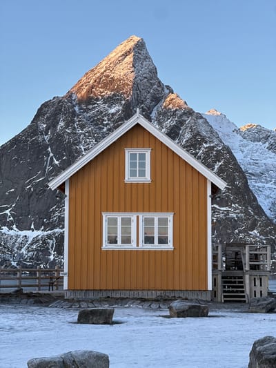 The image displays a prominent orange-brown wooden house situated in Hamnøya, Norway, against a backdrop of rugged, snow-dusted mountains under a clear blue sky. The single-story house features a pitched roof with white trim and a white-framed gable window centrally positioned above two larger, horizontally oriented multi-pane windows on its front facade. Additional white trim lines the house's corners and eaves. A dark-toned foundation is visible beneath the vertical wooden siding. To the immediate right of the structure, a partial view of a wooden deck or stair system is present. A low, light-colored wooden fence runs along the left side of the property, displaying a sign with the text "PRIVAT". The foreground consists of ground covered in a layer of bluish-white snow or ice, interspersed with several dark, rounded rocks. The dominant mountain peak directly behind the house exhibits exposed dark rock and snow, with its upper portion illuminated by a golden sunlight hue. No individuals, animals, or vehicles are visible in the scene.