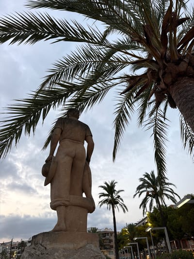 This is a low-angle shot of a stone statue of a man in Port D'alcúdia, Spain. The statue is positioned on a rocky pedestal and is viewed from behind, with the man holding a hat in his left hand and what appears to be a fishing net or sack in his right. He wears a simple tunic and trousers, with his lower legs wrapped. The statue is framed by the fronds of a large palm tree in the foreground, with its trunk also visible. In the background, several other palm trees are scattered throughout the scene, along with buildings that suggest an urban setting. The sky is overcast, with visible clouds. The overall impression is a coastal town atmosphere, possibly near a promenade or public square.