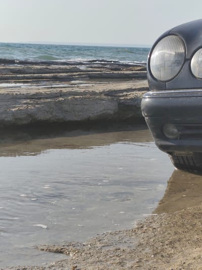 A dark-colored vehicle is partially submerged in shallow water on a rocky shoreline. The front of the car, including headlights and bumper, is visible. The water reflects the sky and the car. In the background, the sea stretches to the horizon under a clear sky. The location is the city of Saiyn, Kazakhstan.