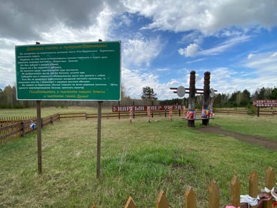 The image shows a natural setting, likely a field or clearing, possibly near Jerdovka, Russia, given the Cyrillic script present on the signs. A large green sign in the foreground contains text in Russian, likely providing information or rules for visitors. Behind this sign, there's a wooden fence enclosing a designated area. Within this area, there are wooden posts or structures adorned with colorful ribbons and cloth, and a bench. The background features trees and a blue sky with scattered clouds. A smaller sign in Cyrillic is also visible in the distance. Overall, the scene suggests a cultural or spiritual site, likely a place of respect or worship for the local population.