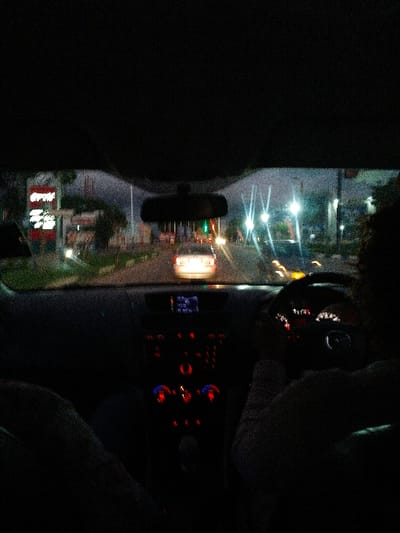 The image is taken from inside a car at night, facing toward the road. Out the windshield, streetlights and building signs are visible. The car's dashboard is visible, illuminated with the instrument panel lights and climate control buttons. A rearview mirror is positioned in the center of the windshield. The driver's hands are visible on the steering wheel. The background is quite dark.