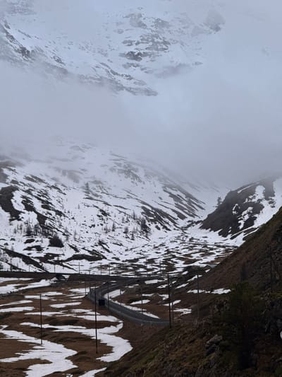The image displays a rugged mountain landscape under heavy fog and overcast skies, characteristic of high alpine regions, possibly near Pontresina, Switzerland. The scene is dominated by steep slopes, patches of snow, and a visible railway line.
In the mid-ground, a railway track runs horizontally across the frame, traversing a dark, rocky mountain face. The track appears to be part of a mountain railway, featuring overhead line supports or protective structures that extend along its path and partially disappear into a dark opening, possibly a tunnel entrance or avalanche gallery, on the right side. The ground surrounding the tracks is a mix of exposed dark rock, earth, and scattered patches of white snow.
Towards the left and background of the image, the mountain peaks are heavily shrouded in thick, grey fog and low clouds. The mist obscures much of the detail, rendering the distant landscape hazy and ethereal, with only faint outlines and large areas of snow-covered terrain discernible.
The overall color palette is muted, featuring various shades of grey from the fog, whites from the snow, and dark browns and blacks from the exposed rock and earth. The lighting is soft and diffused due to the overcast conditions, with no direct sunlight or sharp shadows. There are no people or visible text in the image. The atmosphere is stark and serene, conveying the grandeur and sometimes challenging conditions of a mountainous environment.