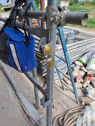 The image depicts a construction site in Settimo Torinese, Italy, featuring scaffolding and building materials. In the foreground, a blue insulated bag and a pair of round-lensed sunglasses are hanging from a metal scaffolding pole. The bag is dark blue with white and grey stripes and a black zipper. The sunglasses have gold-colored frames and yellow lenses. Behind the scaffolding, stacks of metal scaffolding frames are visible, along with piles of bricks and a bag of building material, possibly cement or lime. The ground is covered in dirt and dust, with a length of rope coiled on the right side. A small building with a red-tiled roof and green vegetation is visible in the background. The scene appears to be an outdoor work area under daylight.