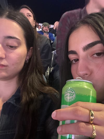 The image captures a dim-lit scene from within a stadium or arena audience in East Rutherford, United States. In the immediate foreground, a person on the right is seen drinking from an open, green aluminum can branded "Goose Island," held in their right hand. Two gold-colored rings are visible on the fingers holding the can: one a plain band, the other intricately patterned. Their dark hair frames their face, which is partially obscured by the can. To the left, a second person with long dark hair is visible from the left side of their face, looking downward or to the left, wearing a dark zippered jacket and a silver hoop earring. In the background, a man with a dark green beanie and a beard is visible, holding a mobile phone, seated amidst other partially visible spectators. Another individual's back and shoulders are visible to the right of the man in the beanie, wearing a red and black plaid shirt. The environment appears to be an indoor or covered outdoor venue with tiered seating.