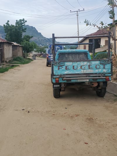 The image depicts a dusty, rural street scene in Ampan, Nigeria. The primary subject is a weathered, turquoise-blue Peugeot pickup truck, viewed from the rear. The truck has a utilitarian design, with a sturdy metal cargo bed and prominent "PEUGEOT" lettering visible on its side. It appears to be parked on a dirt road, with a sparse scattering of small red and dark-colored debris on the ground.

In the background, several buildings with simple construction are visible, some with corrugated metal roofs. Lush green trees and vegetation dot the landscape, and a rugged, rocky hillside rises in the distance under an overcast sky, suggesting a potentially humid or cloudy day. Electrical wires and a utility pole are also present, indicating some level of infrastructure in the area. Another vehicle, partially obscured, is parked behind the Peugeot. A motorcycle is parked on the side of the road near some of the buildings. The overall impression is one of a typical day in a non-urban environment. There are no people actively engaged in activity in the frame.