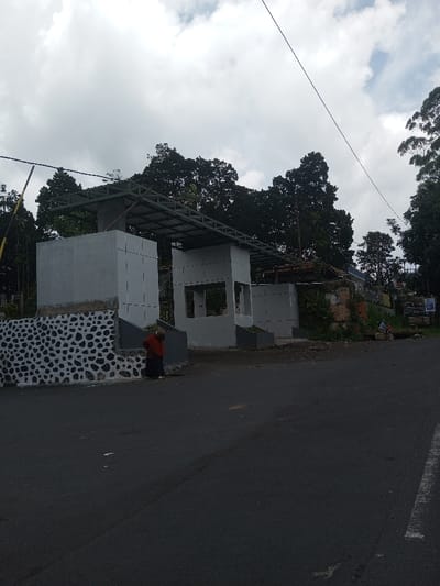 The image depicts a roadside scene in Ura-Dozhi, Bhutan, under an overcast sky. In the foreground, a dark asphalt road, slightly marked with tire impressions, occupies the lower portion of the frame. To the left, a multi-part structure appears to be under construction or renovation, featuring light gray concrete walls and an unfinished metal framework roof. The base of the leftmost section of this structure is adorned with a decorative pattern of dark gray and white stones.
A single individual, dressed in a dark garment with a reddish-brown upper covering, stands near the base of the leftmost structure, facing away from the camera. Further to the right, behind the central portion of the structure, an older building with a tiled roof is partially visible. Adjacent to this, in the far right background, several indistinct figures are seated or standing near what appears to be a small roadside vendor or stall, identifiable by signs and some objects on the ground.
The background is dominated by dense, dark green trees and foliage, suggesting a heavily vegetated area. Overhead, utility lines traverse the sky diagonally from left to right. The overall impression is of a semi-urban or rural roadside area undergoing development.