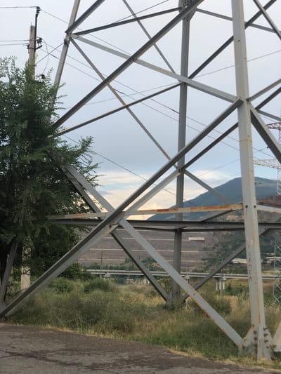 The image shows a close-up view of a large metal power line pylon. The pylon is constructed from grey metal beams, bolted together, with diagonal bracing forming a complex geometric pattern. Some of the metal shows signs of rust, particularly on horizontal beams. To the left of the pylon, a lush green tree with dense foliage partially obscures the structure.
In the background, beyond the pylon's framework, is a landscape that includes a cloudy sky with hints of soft light, suggesting either dawn or dusk. Below the sky, a series of hills or mountains are visible. One prominent hill, covered in green vegetation and with visible terraced areas, dominates the middle ground. Further in the distance, what appears to be a highway or a series of bridges can be seen crossing a valley. There are also signs of industrial activity with some structures and cleared land on the hillsides. The foreground consists of a grey, textured surface that could be asphalt or concrete, and an area of overgrown dry grass and greenery around the base of the pylon. The image appears to be taken in an outdoor, possibly semi-rural or industrial area, given the presence of the power pylon and the distant infrastructure on the hillsides. The location is specified as Khujakent, Uzbekistan.