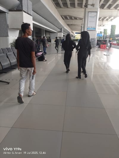 This image captures a daytime scene within a pedestrian concourse at Soekarno-Hatta International Airport (CGK) in the Special Capital Region of Jakarta, Indonesia. The flooring consists of large square tiles in alternating light grey and beige tones, with a darker grey strip defining a central pathway. On the left, a row of dark metal-framed waiting benches is positioned. A male, viewed from the back, wearing a dark t-shirt, light-colored trousers, and sandals, walks towards the left foreground, holding a small rectangular object in his right hand. In the mid-ground, two females dressed in black professional attire (jackets and trousers) walk away from the viewer towards the right; the individual on the far right wears a black hijab and carries white papers in her left hand. Structural components include large concrete pillars supporting an overhead beam system. In the background right, a vertical pillar displays a "Garuda Indonesia" advertisement. Beyond this, green foliage and red barriers are visible, indicating an adjacent outdoor area likely used for vehicle access. Several other blurred figures are present in the background, either walking or seated.
