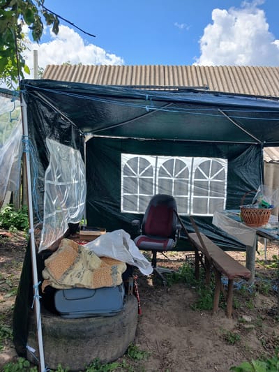 The image depicts an outdoor scene, possibly in Hlushky, Ukraine. It features a makeshift shelter constructed from a dark green tarp, supported by poles. The shelter has at least two windows covered with white material featuring a grid pattern. Inside, a red and gray office chair is visible. Outside the shelter, on the ground, lie various objects, including a large tire, a blue suitcase, a folded blanket with an orange and beige pattern, and a white plastic bag. A woven basket hangs from a structure to the right. The ground is dirt and overgrown with some greenery and plants. In the background, a corrugated roof structure is visible, and above, a blue sky with white clouds suggests it is daytime. The overall impression is one of a rustic, possibly temporary, outdoor setup. There is no visible text in the image.