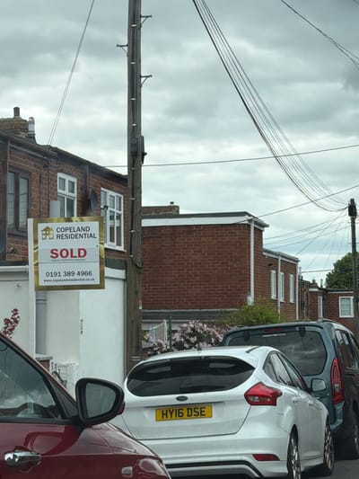 The image shows a residential street in the city of Durham, United Kingdom. A "SOLD" sign from Copeland Residential is visible in front of a brick building. A red car is in the foreground, with a white car and a grey van parked behind it. Overhead, there are telephone wires and a wooden utility pole. The sky is overcast. The cars partially obscure the view of the houses along the street.