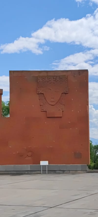 The image shows a large, rectangular monument in Araks, Armenia. The monument is made of reddish-brown stone and features a carved face near the top center. Below the face, on the monument's surface, are two more carvings. The monument is situated on a concrete platform, and in front of the monument is a small, white sign or marker. The background consists of a blue sky with scattered white clouds, and there is some greenery visible on either side of the monument, suggesting the presence of trees or foliage.
