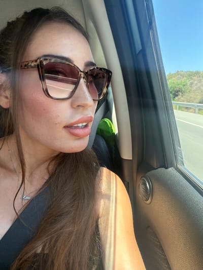 The media file is a portrait-oriented close-up shot of a young woman inside a car. The woman is wearing large, tortoise-shell sunglasses and has her mouth slightly open, revealing her teeth. She has on lipstick and her hair, which is long and brown, is falling around her. A thin silver necklace with a pendant is visible on her neck. The background shows the interior of a car, including the car door panel, a speaker, and a window with a view of a road and vegetation outside, suggesting a sunny day. The light indicates it is daytime, and the scene is likely set in a warm climate. There is no visible text in the media file. The overall impression is a casual, self-taken photograph, likely a selfie, capturing a moment during a car ride.