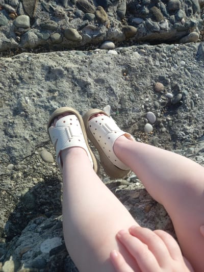 The image shows a close-up of a child's legs and feet, wearing light-colored sandals, resting on a rocky surface. The child's skin appears fair and smooth. The sandals are made of a flexible material and have a practical design with straps and perforated details. They are worn over bare feet. The rocky ground is composed of various stones, pebbles, and some larger, flatter rocks, suggesting an outdoor setting. The lighting indicates it is daytime, likely sunny, casting shadows and highlighting the textures of the rocks and the child's skin. The scene appears peaceful and relaxed, with the child's legs casually placed. The location context provided is Gwandra, Abkhazia, which implies a coastal or riverine environment given the type of rocks. There is no visible text in the image.