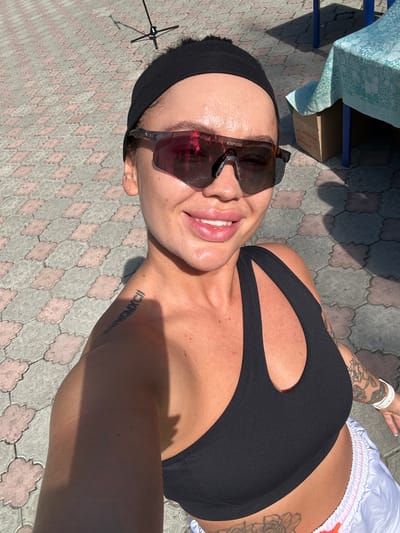 A woman is taking a selfie outdoors on a sunny day. She is wearing a black headband, sunglasses, and a black sports bra. She has a tattoo on her left shoulder blade that appears to read "MC MCII" in Roman numerals, and other tattoos on her arm and torso. She is smiling and her skin is glistening with sweat. The background shows a paved area with a repeating cloud-like pattern, a table covered with a blue and white patterned cloth, and some boxes. There is also a shadow from a pole or stand. The scene appears to be in Duvankul', Russia.