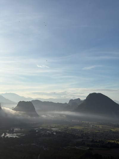 An aerial view captures a misty mountain landscape near Naka, Laos, bathed in the soft light of early morning or late afternoon. Silhouetted against a pale blue sky with wispy clouds are several jagged mountain peaks, some veiled in ethereal white mist that drifts through the valleys. The mist obscures the lower portions of some mountains, creating a dramatic, layered effect.

Below the misty peaks, a verdant valley unfolds, featuring a patchwork of agricultural fields. Some fields appear to be flooded, reflecting the sky, while others are dry and cultivated. Small clusters of buildings, suggesting a village or settlement, are scattered throughout the valley, interspersed with trees and vegetation. A winding road is faintly visible in the lower foreground.

The sky is populated by several small, dark shapes that appear to be birds in flight, adding a sense of life and movement to the serene scene. The overall atmosphere is tranquil and majestic, highlighting the natural beauty of the mountainous terrain.