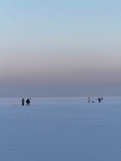 The image displays a vast, flat, snow-covered landscape, characteristic of a frozen body of water, situated in Liutizh, Ukraine. The sky transitions from a pale blue overhead to a muted purple-grey at the distant horizon, indicating ambient light conditions. Six human figures are observed across the mid-ground.

On the left, two individuals stand on the frozen surface. One wears a light-colored jacket and dark pants, facing slightly right. The second, in a dark reddish jacket and dark pants, faces forward. A small, light-colored object rests on the snow to their left. Faint linear impressions are visible on the snow behind them.

On the right, a group of three individuals is clustered. The leftmost wears a light jacket and dark pants, facing left, with a dark object on the snow nearby. A central figure, in a dark jacket and dark pants, stands with arms extended horizontally. A third person, in a light jacket, is positioned further back and to the right within this cluster. A sixth, indistinct figure appears as a dark silhouette further right on the horizon line.

The individuals are spaced out across the expansive, uniform frozen surface. Their primary action consists of standing. Specific fishing gear or active fishing is not clearly discernible, though the presence of objects on the ice near the groups could be consistent with ice fishing equipment.