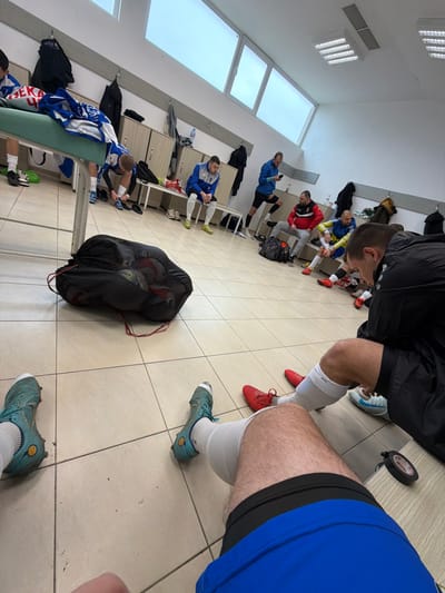 An indoor scene depicts a men's changing room, likely within a sports facility in Suvorovo, Bulgaria. The room features light-colored tiled flooring, white walls, and a white ceiling with rectangular fluorescent light fixtures and vents. A series of light-brown wooden lockers with open compartments and benches extend along the back wall, below a row of large, horizontally oriented windows providing natural light.

Eight adult males are present, mostly in athletic attire. In the immediate foreground, the perspective is from a seated individual, showing their legs in blue shorts, white athletic socks, and teal-colored soccer cleats. To their right, another pair of legs with white socks and red soccer cleats is visible, alongside a black stopwatch with a red button.

Further back, a man in a blue and white uniform is leaning forward, adjusting or tying his shoes near a green-topped bench or table. Another man in a similar blue and white uniform sits on a wooden bench. To his right, a man in a blue jacket and dark shorts stands with one leg elevated on a bench, looking at a smartphone. A man in a red and black jacket and grey pants is seated on a bench, with a black duffel bag near his feet. Adjacent to him, a man in a yellow and blue athletic shirt is also seated, appearing to adjust his red cleats. A figure in a black jacket is partially visible on the far right, seated.

A black mesh bag, containing multiple pairs of athletic shoes, rests on the tiled floor in the mid-foreground. Various items of clothing, bottles, and personal shoes are scattered on the benches and locker shelves.