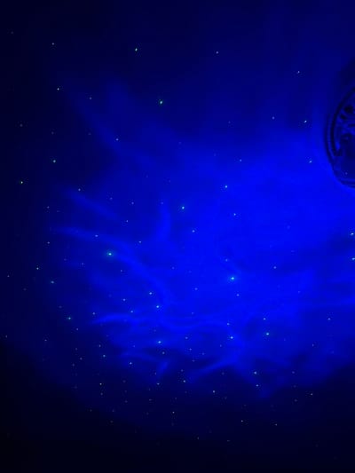 The image depicts a dark blue ceiling with numerous small, glowing green points of light, resembling stars. There are also faint, wispy patterns of brighter blue light, suggesting a nebula or celestial display. In the upper right corner, a portion of a decorative molding is visible, which appears to be circular and ornate. The overall impression is that of a simulated night sky. The location in Komsomolskiy, Russia, is not discernible from the visual information presented in the image. There are no people, animals, or discernible actions taking place. The scene is static and focuses solely on the visual effect of the illuminated ceiling.