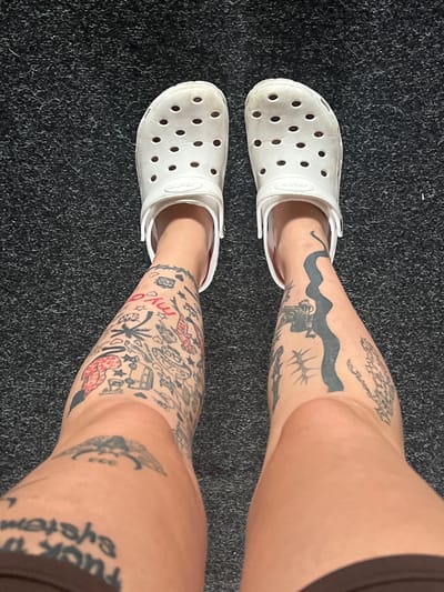 The image shows a top-down view of a person's legs wearing white Crocs, standing on a dark, textured carpet. The legs are covered in various black and red tattoos. The left leg features a collection of smaller, cartoon-like designs, including fish, stars, and what appears to be a rocket. The right leg has a larger, wavy black line tattoo running vertically, with other designs around it, including a creature resembling a gargoyle. The person is wearing dark brown shorts that are visible at the bottom of the frame. The overall setting appears to be indoors, on a carpeted surface.