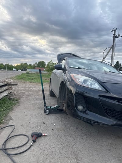 The image shows a black Mazda car being serviced on a concrete surface. The front driver-side wheel is removed, and the car is raised with a green and black hydraulic jack. Scattered on the concrete are several lug nuts and a red and black impact wrench connected to a black air hose. The car's hood is open. The setting appears to be outdoors, possibly on a roadside in Druzhino, Russia. A utility pole with wires is visible in the background, and the sky is overcast. In the distance, there are trees, buildings, and other vehicles. There is no indication of the presence of people.